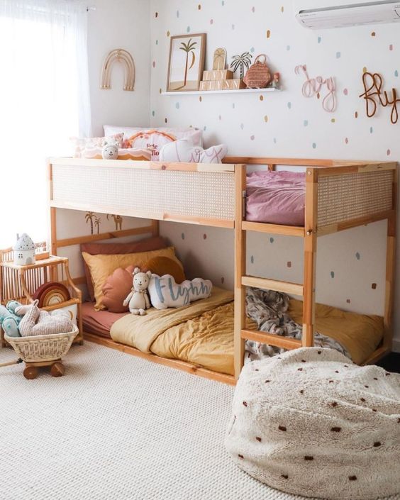 a bright and cool shared girls' bedroom with a bunk bed with warm-colored bedding, a pouf with pompoms, shelves fro storage and calligraphy