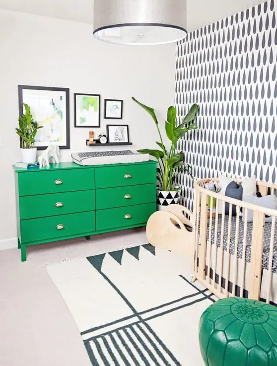 a bright mid-century modern meets boho nursery with an accent wall, an emerald green dresser, a stained crib, a green pouf, potted plants and a gallery wall