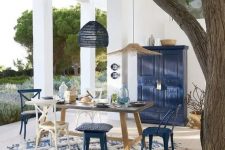 a bright seaside patio in blue and white, with a wooden table and white and navy chairs and stools, pendant lamps and a navy sideboard