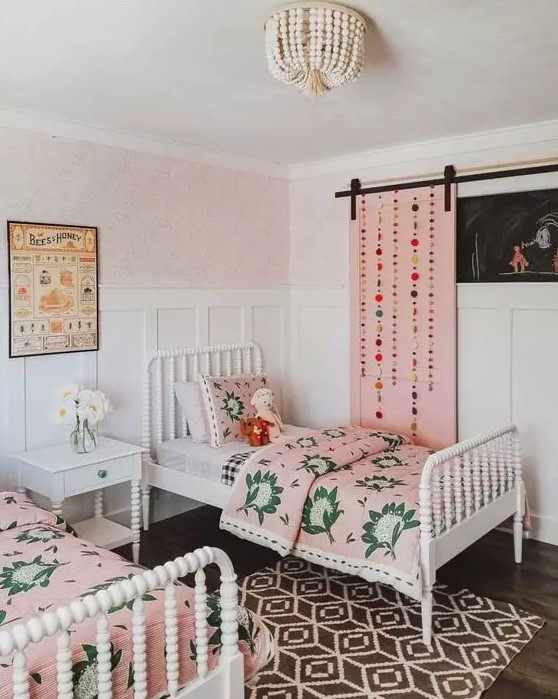 a bright shared girls' bedroom with white vintage furniture, pink bedding and a pink barn door with pompoms, a printed rug