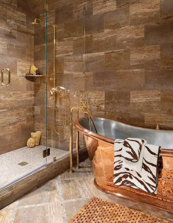 a catchy bathroom clad with brown stone tiles, a copper bathtub and copper and gold fixtures is pure chic