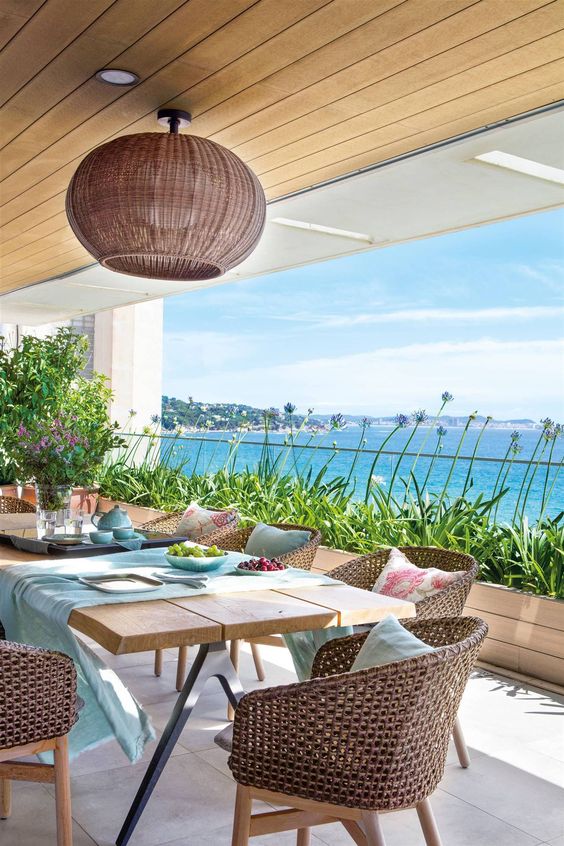 a cheerful coastal terrace with a wooden trestle dining table, woven chairs, a long planter with greenery and blooms and a jaw dropping view