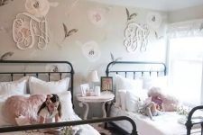 a chic farmhouse shared girls’ bedroom with a floral accent wall and monograms, with metal beds and white and pink bedding, a round table as a nightstand