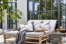 a chic modern country terrace with a wooden deck, rattan furniture, small side tables, lots of greenery around and lots of pillows