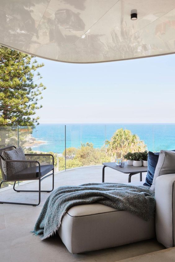 a coastal terrace with a neutral low sofa, a grey chair, a large coffee table and some potted greenery plus a sea view
