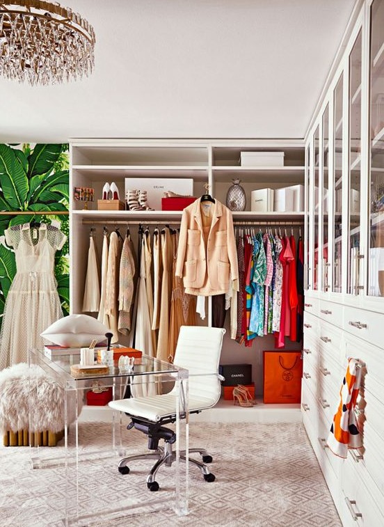 a colorful glam cloffice with a crystal chandelier, a printed rug, a makeshift closet, a largre closed storage unit, an acrylic desk and a white chair