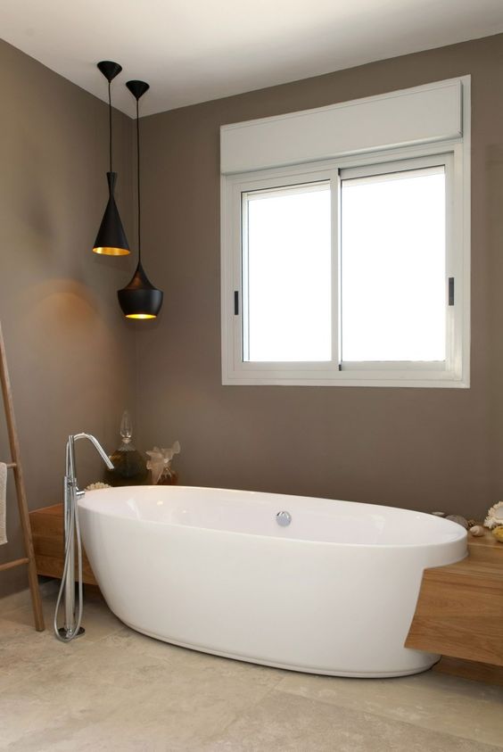 a contemporary brown bathroom with a neutral stone tile floor, an oval tub, black pendant lamps