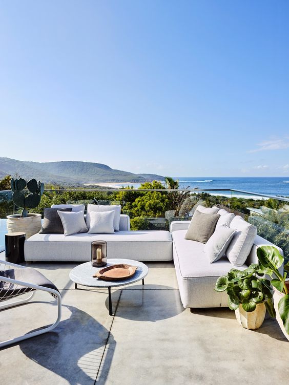 a contemporary coastal terrace with low neutral sofas and pillows, a low coffee table and a leather chair, lots of greenery and a sea view