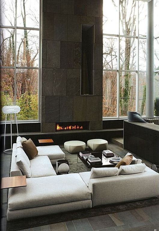 a contemporary living room with floor to ceiling windows, a panel with a built-in fireplace, a low neutral sectional and some poufs is amazing