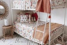 a cool boho shared girls’ bedroom with a white metal bunk bed, floral print bedding, a printed rug, a rattan chair and a nightstand, some art