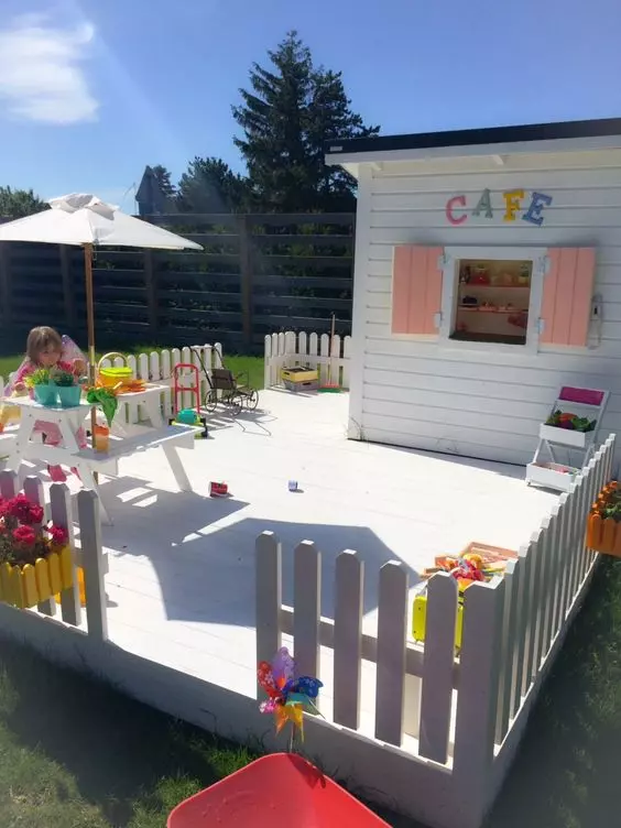 a cool kids' play space with a mini cafe, a wooden dining set with an umbrella, some potted plants and blooms