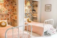 a cool shared girls’ bedroom with niches done with floral wallpaper and shelves, white metal beds, little play cribs and pink bedding