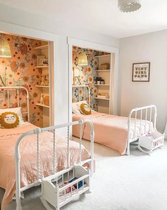 a cool shared girls' bedroom with niches done with floral wallpaper and shelves, white metal beds, little play cribs and pink bedding