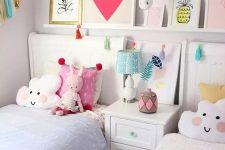 a cute and bright shared girls’ bedroom with white furniture, pastel bedding, a ledge gallery wall, a banner and colorful toys and pillows