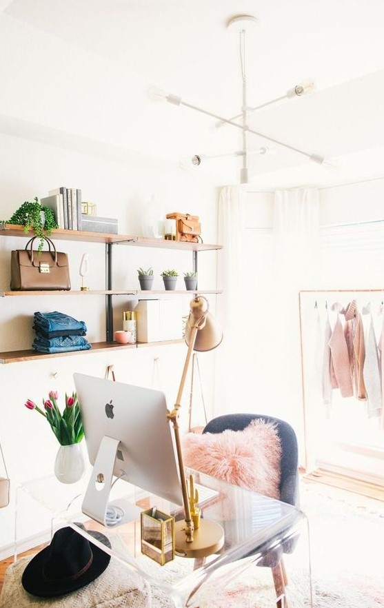 a cute and glam cloffice with open shelving, a makeshift closet, a white chandelier, an acrylic desk, a grey chair and touches of pink