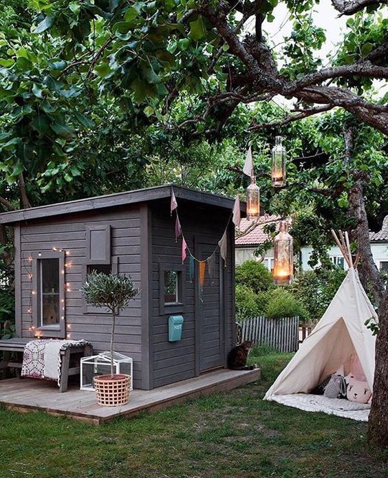 a cute and stylish black modern kids' playhouse with lights and a banner, a small porch and a bench outside plus a teepee in front of it