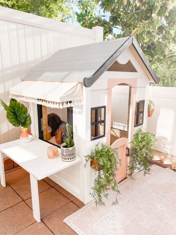 a cute kids' playhouse in white and pink, with potted greenery, a table is a great idea for a backyard