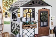 a cute outdoor kids’ playhouse with a dark roof, with potted greenery and a wreath, some decor and lanterns is amazing