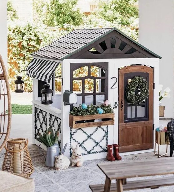 a cute outdoor kids' playhouse with a dark roof, with potted greenery and a wreath, some decor and lanterns is amazing