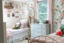 a cute shared girls’ bedroom with a pastel floral wall, white metal beds with pink and white bedding, wall-mounted shelves and toys is very cute