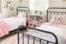 a farmhouse girls’ bedroom with a white planked accent wall, metal beds, a nightstand, pink and blue bedding, blooms and gold sconces
