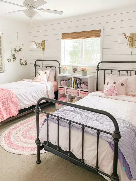 a farmhouse girls' bedroom with a white planked accent wall, metal beds, a nightstand, pink and blue bedding, blooms and gold sconces