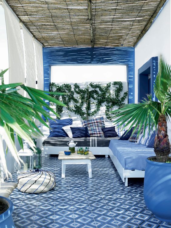a gorgeous seaside terrace with curtains, blue framing, blue tiles on the floor and a corner sofa with blue pillows
