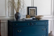 a gorgeous vintage navy dresser with vintage handles, artwork, some branches in a vase, stacked books and a bowl for storage