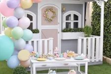 a grey and white kids’ playhouse with potted greenery, a faux bloom wreath, an Easter tablescape and pastel balloons