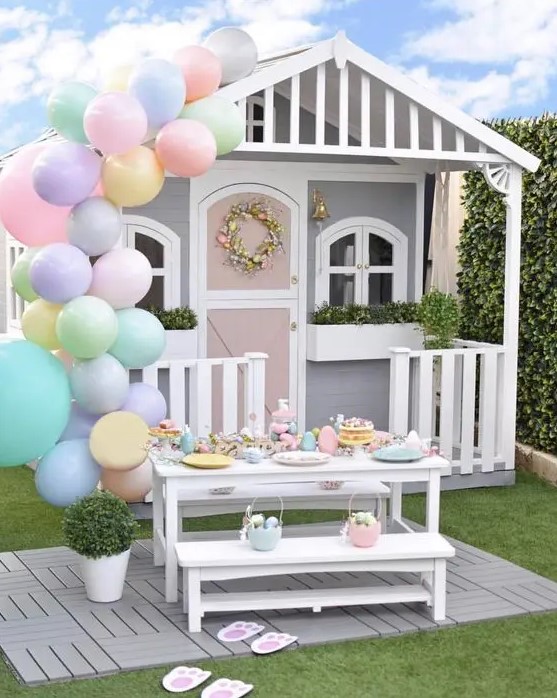 a grey and white kids' playhouse with potted greenery, a faux bloom wreath, an Easter tablescape and pastel balloons