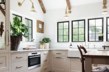 a light-filled barn kitchen with wooden beams, gilded sconces, neutral cabinets, white stone countertops, a table as a kitchen island