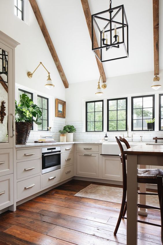 a light-filled barn kitchen with wooden beams, gilded sconces, neutral cabinets, white stone countertops, a table as a kitchen island