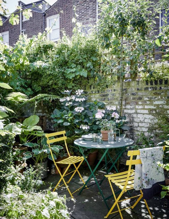 a little garden dining nook by the fence, with blooms and greenery around, a green table and yellow chairs is a lovely space