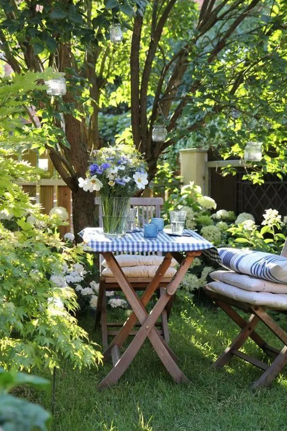 a little garden dining space on a green lawn, under the trees, with hanging candleholders and gold wooden chair plus cool textiles