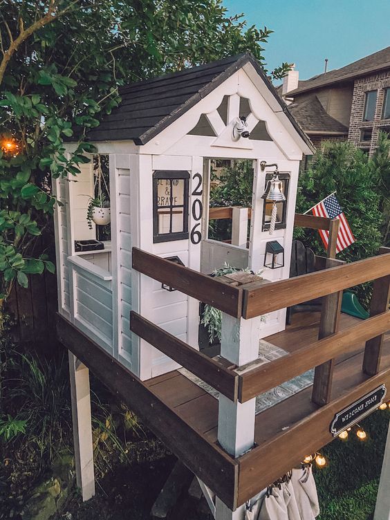a lovely modern kids' playhouse in black and white raised above the ground, with potted plants, lights and a flag