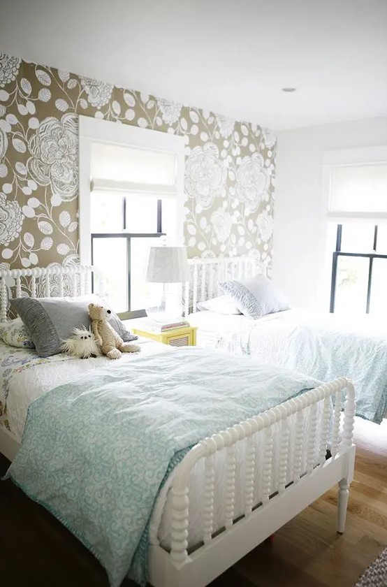 a lovely pastel shared girl bedroom with an accent botanical wall, white wooden beds, blue and grey bedding, a bright nightstand and a table lamp