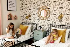 a lovely shared girls’ bedroom with a statement accent wall, black forged beds with neutral bedding, brass sconces and pink house-shaped shelves