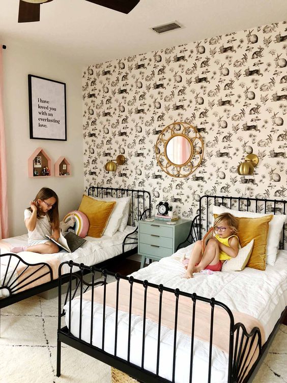 a lovely shared girls' bedroom with a statement accent wall, black forged beds with neutral bedding, brass sconces and pink house-shaped shelves