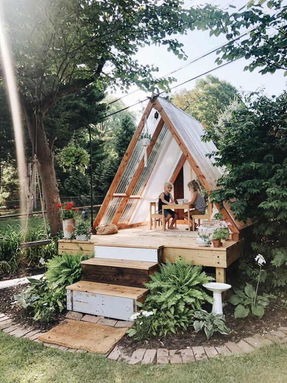 a lovely teepee style playhouse with glass panels, a porch with potted blooms is a cool solution for a boho or modern backyard
