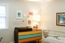 a mid-century modern nursery with a colorful dresser, a white crib with neutral bedding, a black and white printed rug and a gallery wall