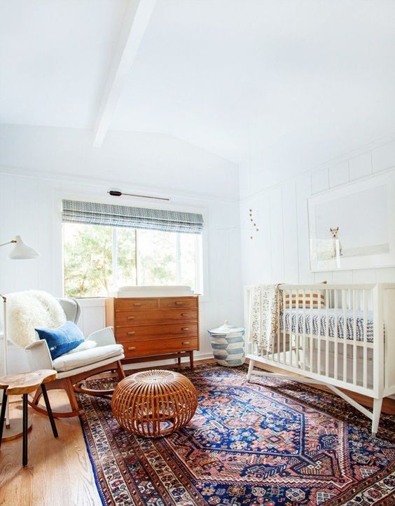a mid-century modern nursery with a neutral crib and printed bedding, a bold printed rug, a rocker chair with a pillow and a stained dresser