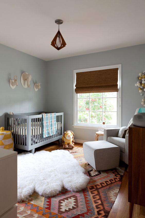 a mid-century modern nursery with light blue walls, a grey crib, layered rugs, a grey chair with a footrest and woven blinds