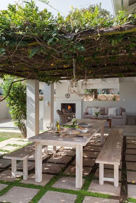 a modern country terrace with a living roof over the space, a whitewashed dining set with benches and an outdoor living room by the fireplace
