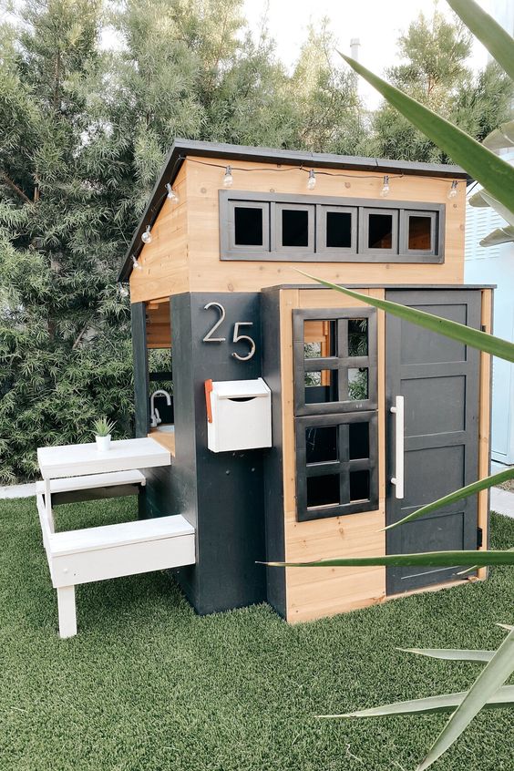 a modern farmhouse kids' playhouse clad with black and stained wood, with windows and a door, a house number and built in benches and a table
