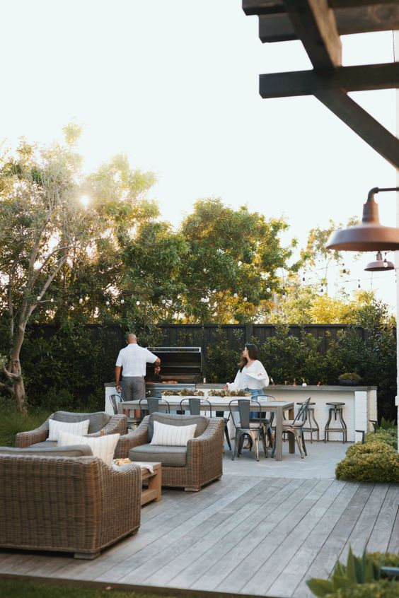 a modern farmhouse terrace done in neutrals, with wicker seating furniture, a wooden table, a table and metal chairs, a bar counter with a grill