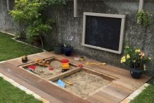 a modern outdoor play space with a sandbox and a deck around it, potted blooms, a chalkboard on the wall