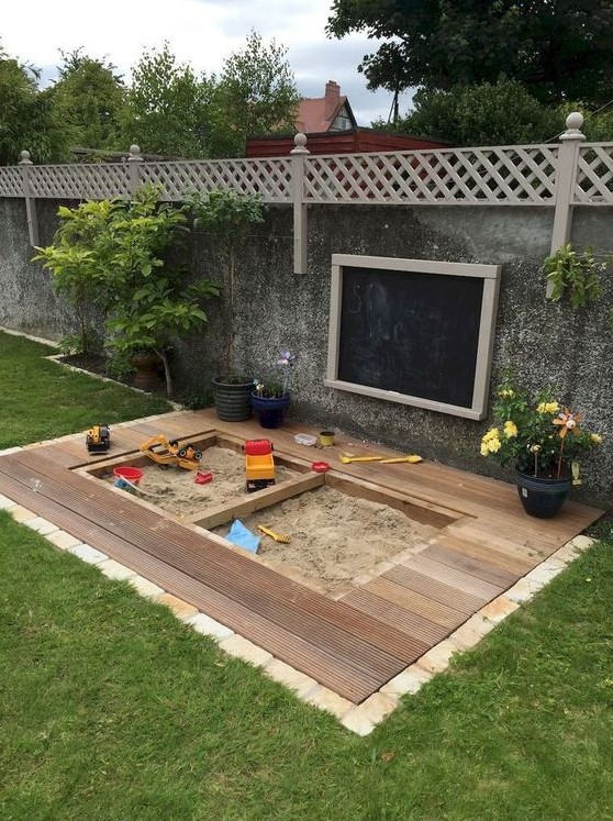 a modern outdoor play space with a sandbox and a deck around it, potted blooms, a chalkboard on the wall
