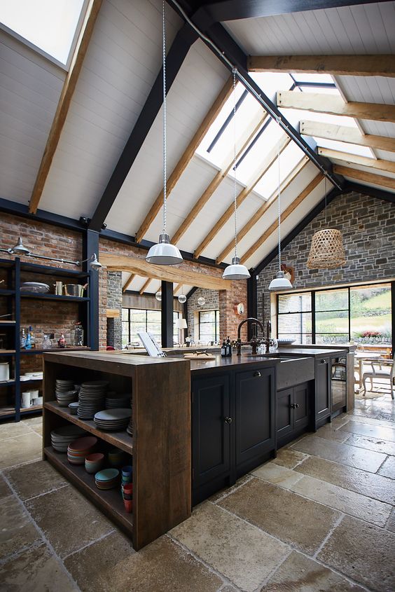 a moody kitchen with skylights, pendant lamps that highlight the double-height ceiling, dark cabinets, a wooden console table and stone walls for a texture