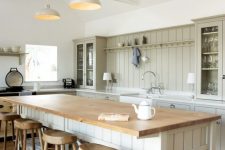 a neutral barn kitchen with light green planked cabinets, a large planked kitchen island with a butcherblock countertop, wooden stools and pendant lamps