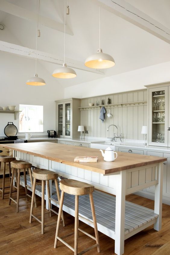 a neutral barn kitchen with light green planked cabinets, a large planked kitchen island with a butcherblock countertop, wooden stools and pendant lamps
